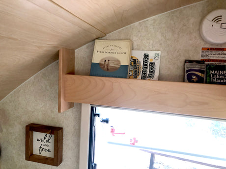 RV rental interior: a light wood shelf with books, games, and a travel guide. A smoke alarm and "wild and free" sign are visible.