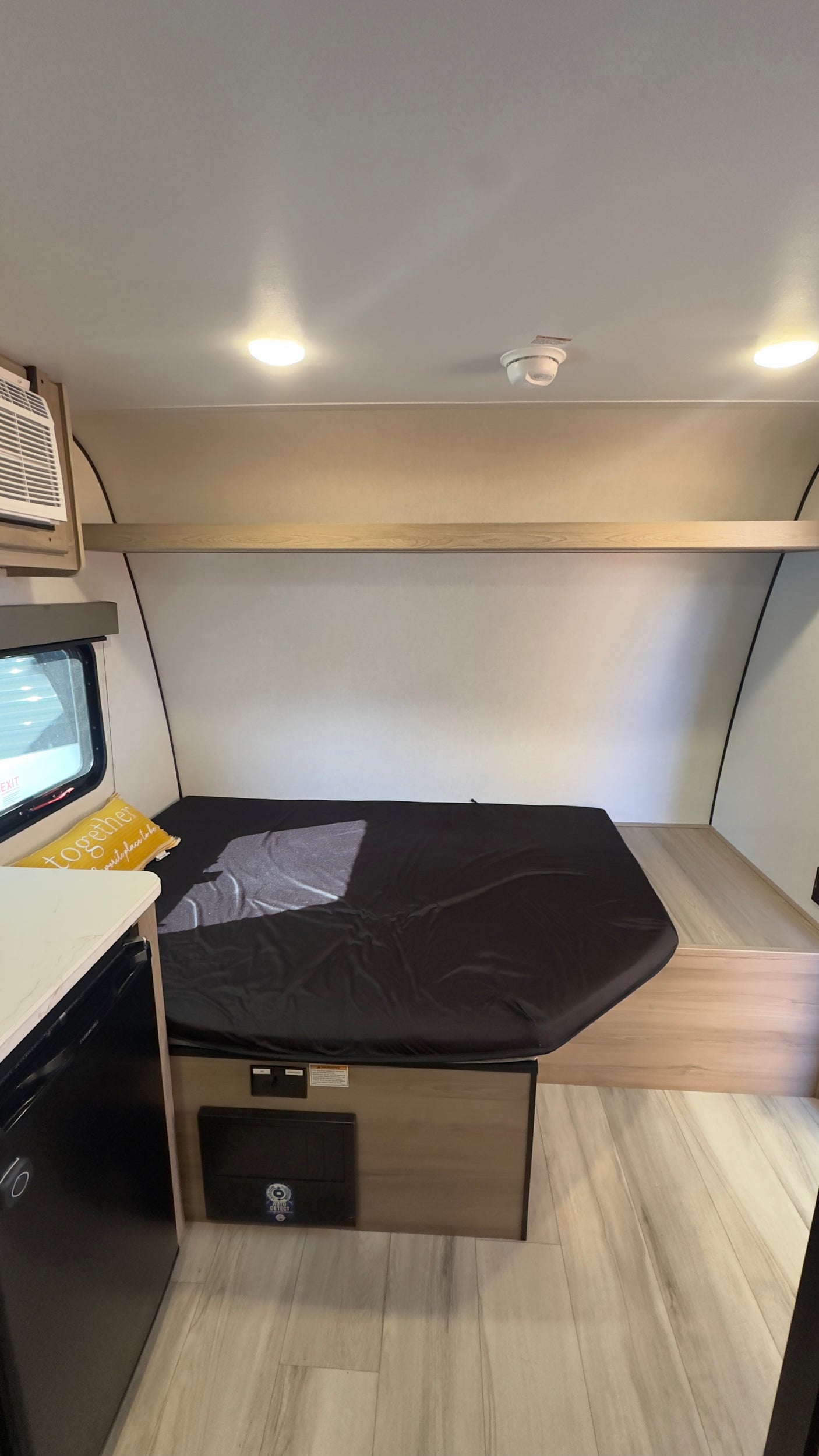 RV rental interior: a compact sleeping area featuring a bed with a black cover, an overhead shelf, and an AC unit.