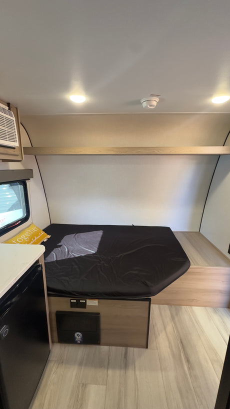 RV rental interior: a compact sleeping area featuring a bed with a black cover, an overhead shelf, and an AC unit.
