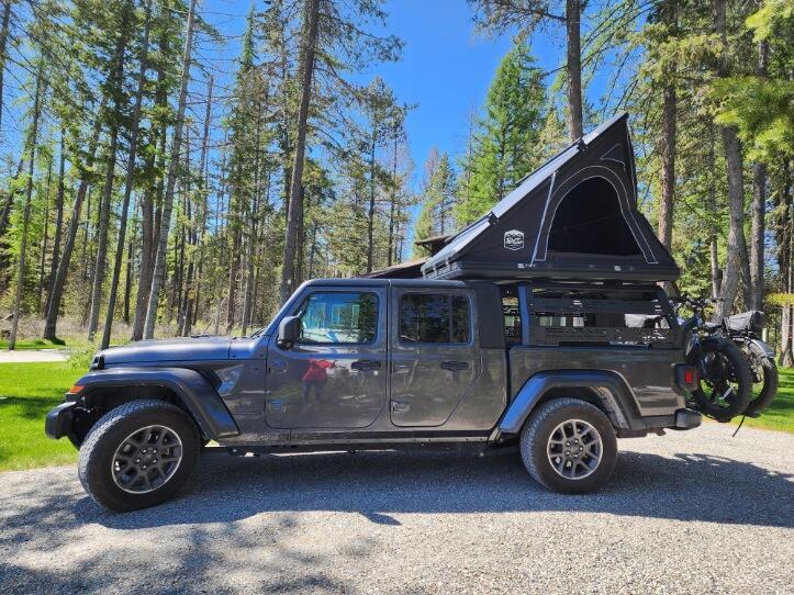 Gray Jeep Gladiator truck with open rooftop tent and bikes, parked in a sunny forest. An ideal RV rental for outdoor adventures. — photo 4