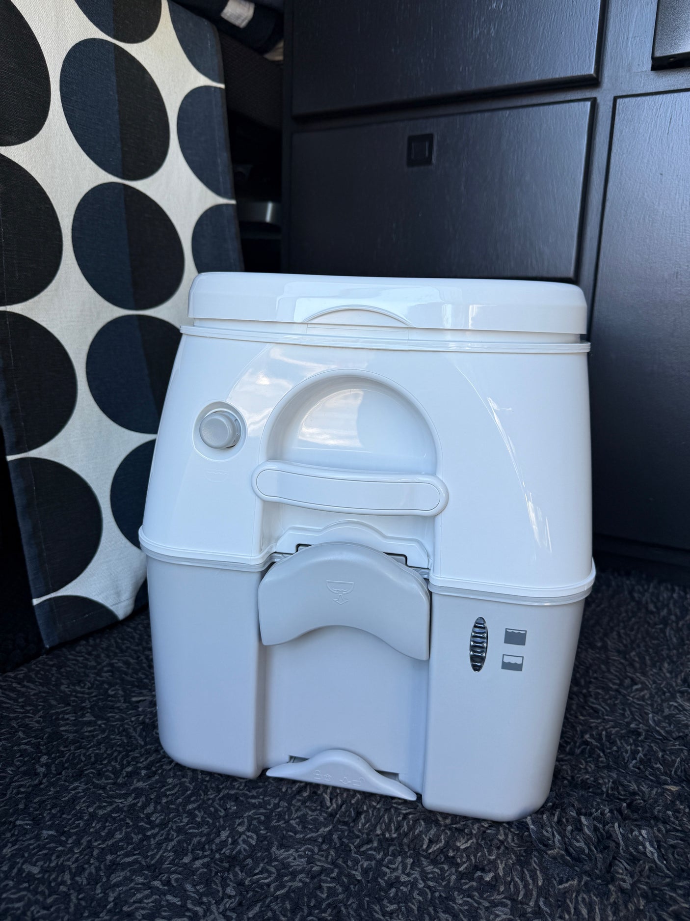 Compact portable toilet, a key feature in many RV rental vehicles, sitting on dark carpet with patterned fabric.
