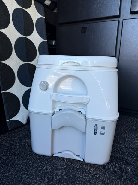 Compact portable toilet, a key feature in many RV rental vehicles, sitting on dark carpet with patterned fabric.