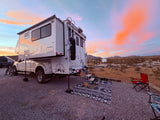 An RV rental, a white truck camper, is set up at a desert campsite under a vibrant sunset sky with an outdoor rug & chairs.