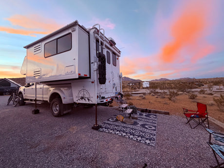 An RV rental, a white truck camper, is set up at a desert campsite under a vibrant sunset sky with an outdoor rug & chairs.