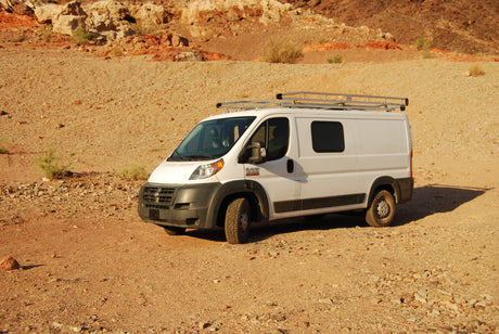 White camper van, an RV rental, with a roof rack on a sunny gravel road in a rugged desert landscape.