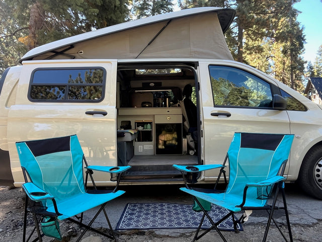 Outdoor living with an RV rental: A beige pop-top camper van, interior visible, flanked by two turquoise camping chairs and an outdoor rug.