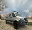 A silver Mercedes-Benz Sprinter camper van for rv rental, parked on gravel under a cloudy, twilight sky.