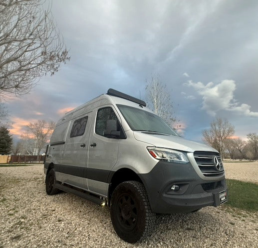 A silver Mercedes-Benz Sprinter camper van for rv rental, parked on gravel under a cloudy, twilight sky.
