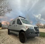 A silver Mercedes-Benz Sprinter camper van for rv rental, parked on gravel under a cloudy, twilight sky.