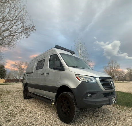 A silver Mercedes-Benz Sprinter camper van for rv rental, parked on gravel under a cloudy, twilight sky.