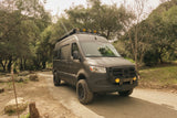 Rugged dark grey off-road Sprinter van rv rental, equipped with custom lights and tires, parked on a dirt path in a wooded area.