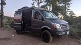 A custom dark gray Mercedes Sprinter van, perfect for an rv rental, parked on a dirt trail in a forest with an open side door.
