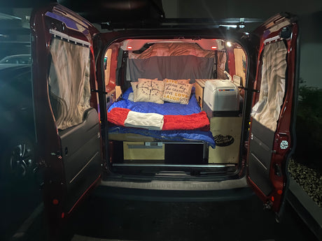 Cozy RV rental camper van interior, featuring a bed with pillows, blankets, and a portable fridge. Ready for van life adventures!