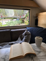 Cozy RV rental: a book and coffee cup on a table, with an open window view of prickly pear cacti & greenery outside.