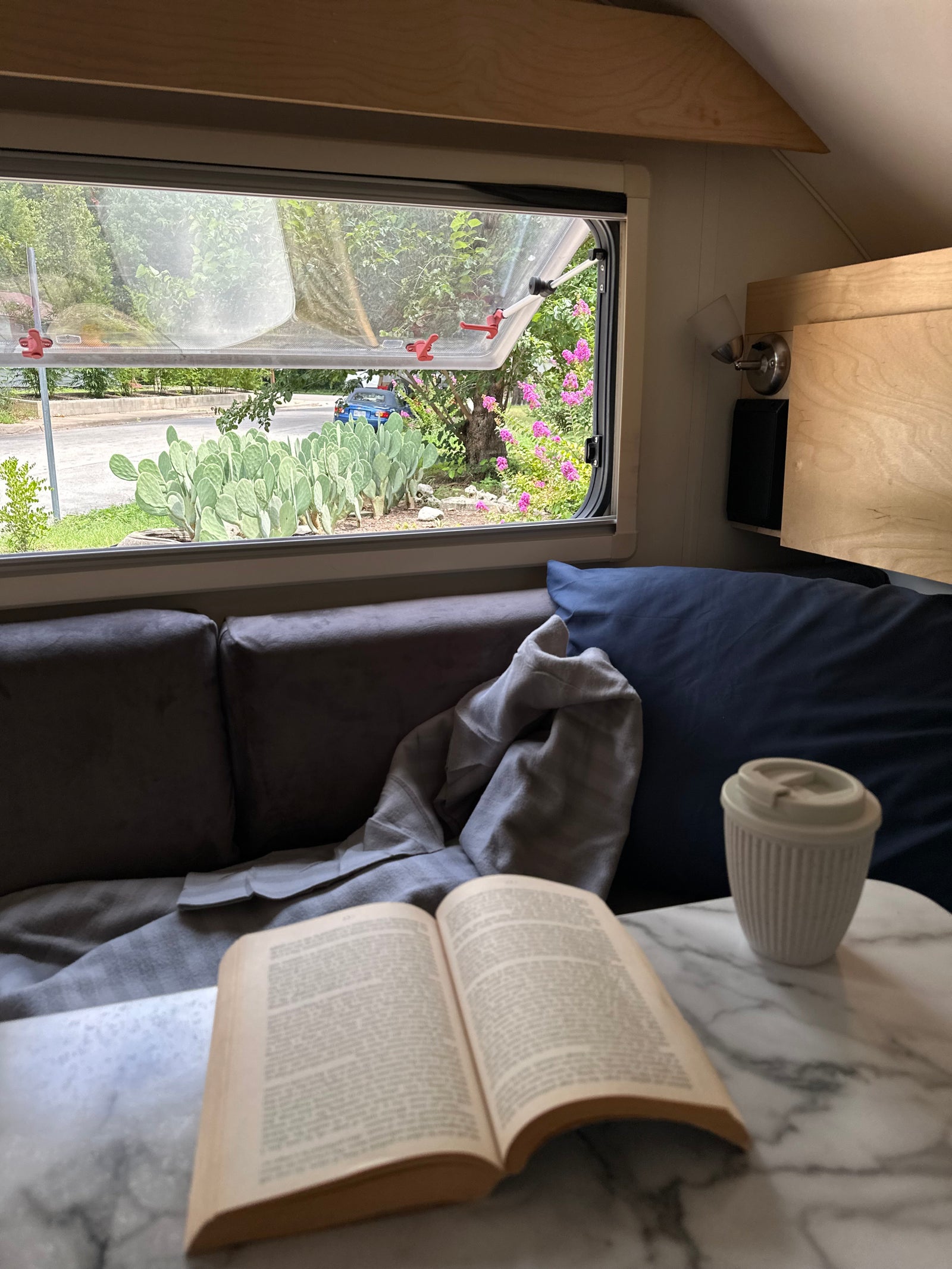 Cozy RV rental: a book and coffee cup on a table, with an open window view of prickly pear cacti & greenery outside.