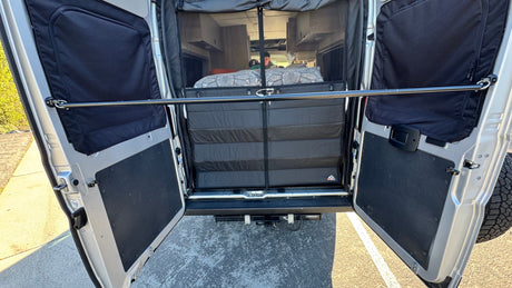 RV rental rear view: open van doors show a functional sleeping setup with a bug screen, insulated panels, and a safety bar.