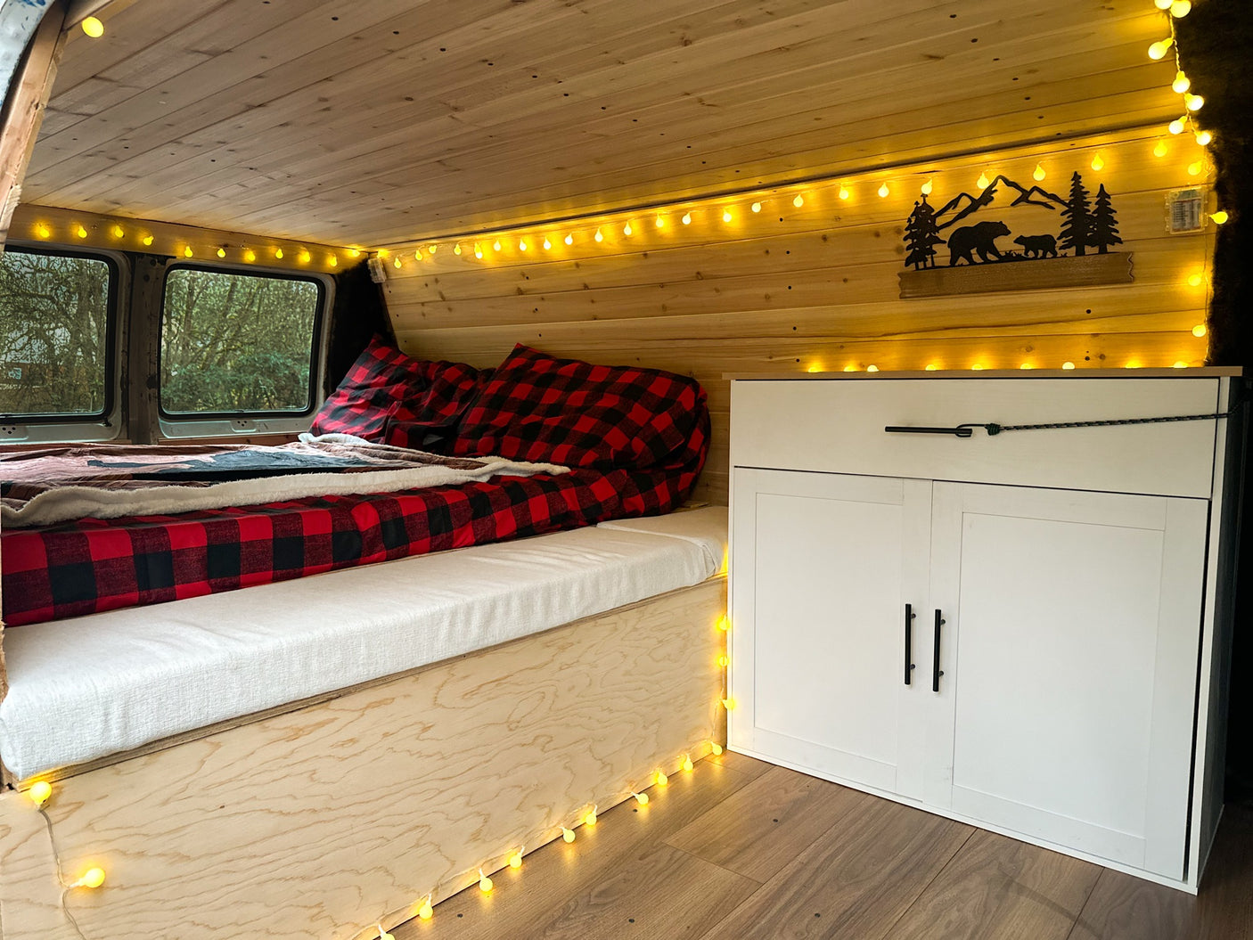 Cozy custom rv rental interior featuring wood paneling, string lights, a plaid bed, and built-in storage cabinets.