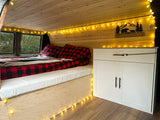 Cozy custom rv rental interior featuring wood paneling, string lights, a plaid bed, and built-in storage cabinets.