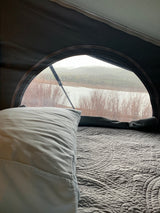 Cozy RV rental bed with a view of a rainy lake and hills. Wiper visible on the arched window, raindrops cover the glass.
