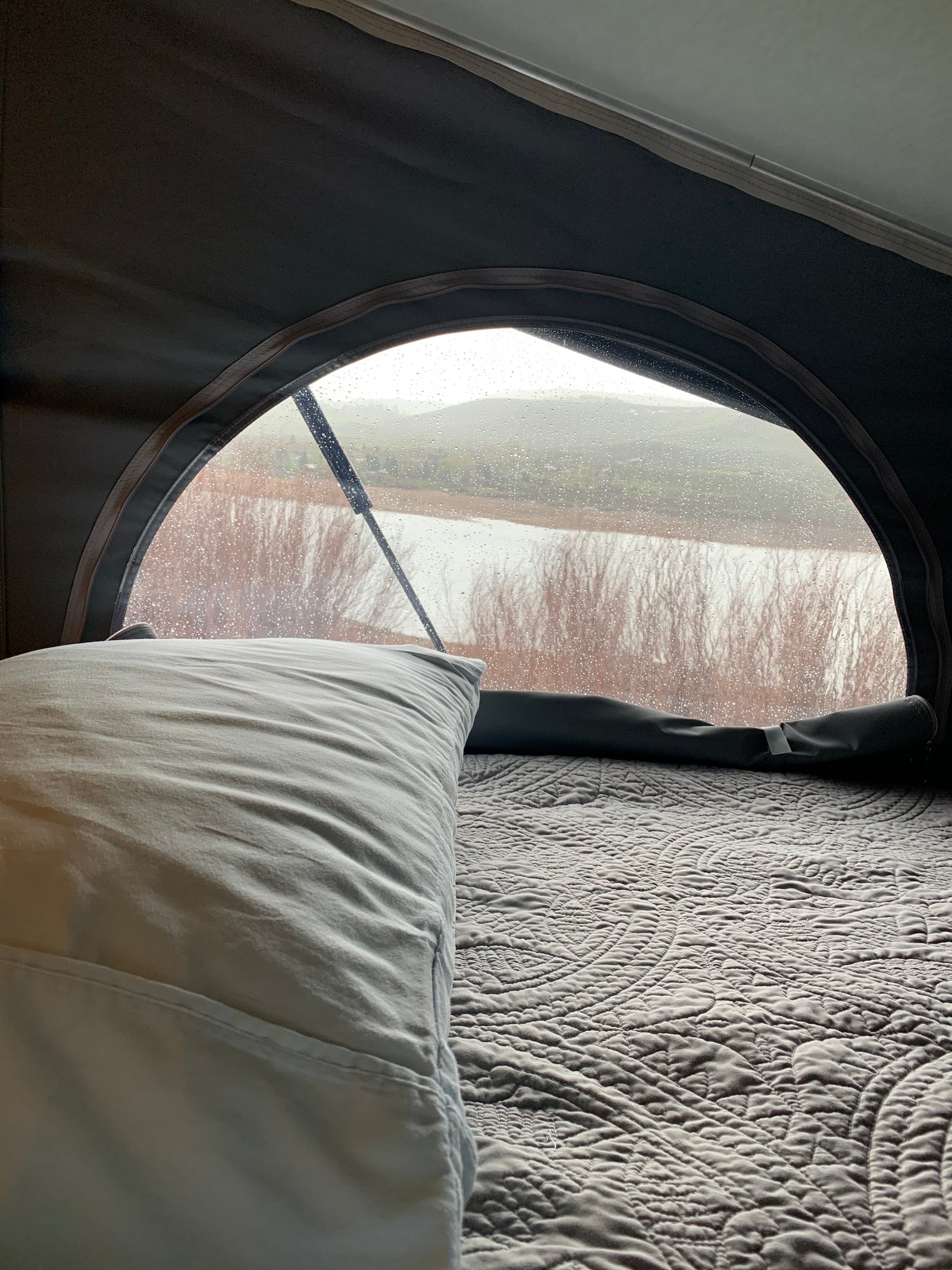 Cozy RV rental bed with a view of a rainy lake and hills. Wiper visible on the arched window, raindrops cover the glass.