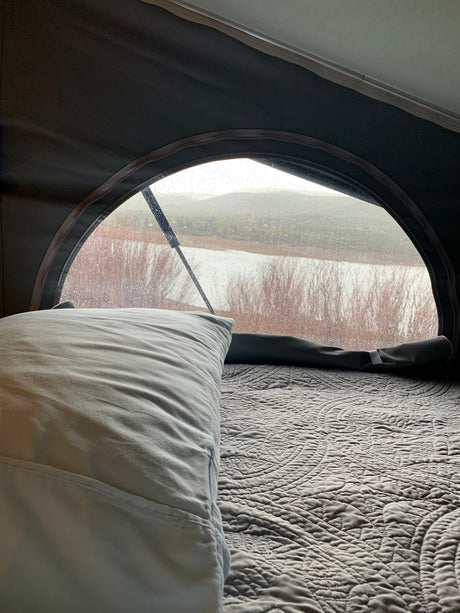 Cozy RV rental bed with a view of a rainy lake and hills. Wiper visible on the arched window, raindrops cover the glass.