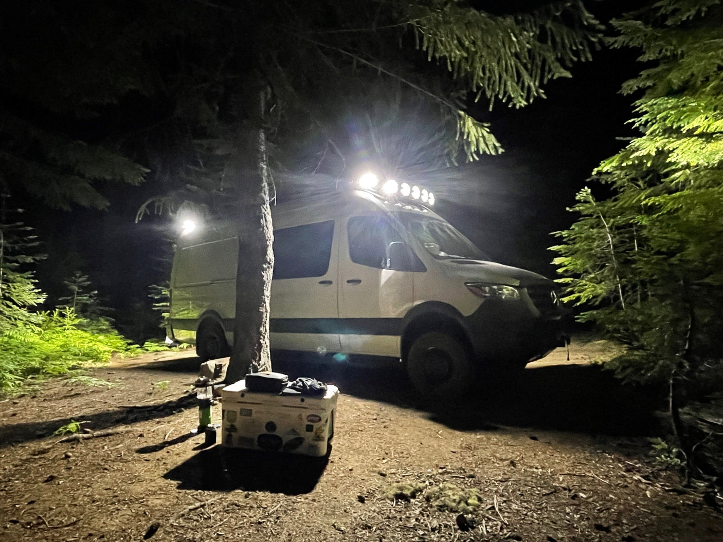 White rv rental van illuminated at night in a dense forest. Camping setup with cooler. Perfect for adventure.