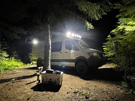 White rv rental van illuminated at night in a dense forest. Camping setup with cooler. Perfect for adventure.