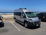Ram ProMaster RV rental parked at a sunny beach. Ocean, mountains, & people under a blue canopy in the background.