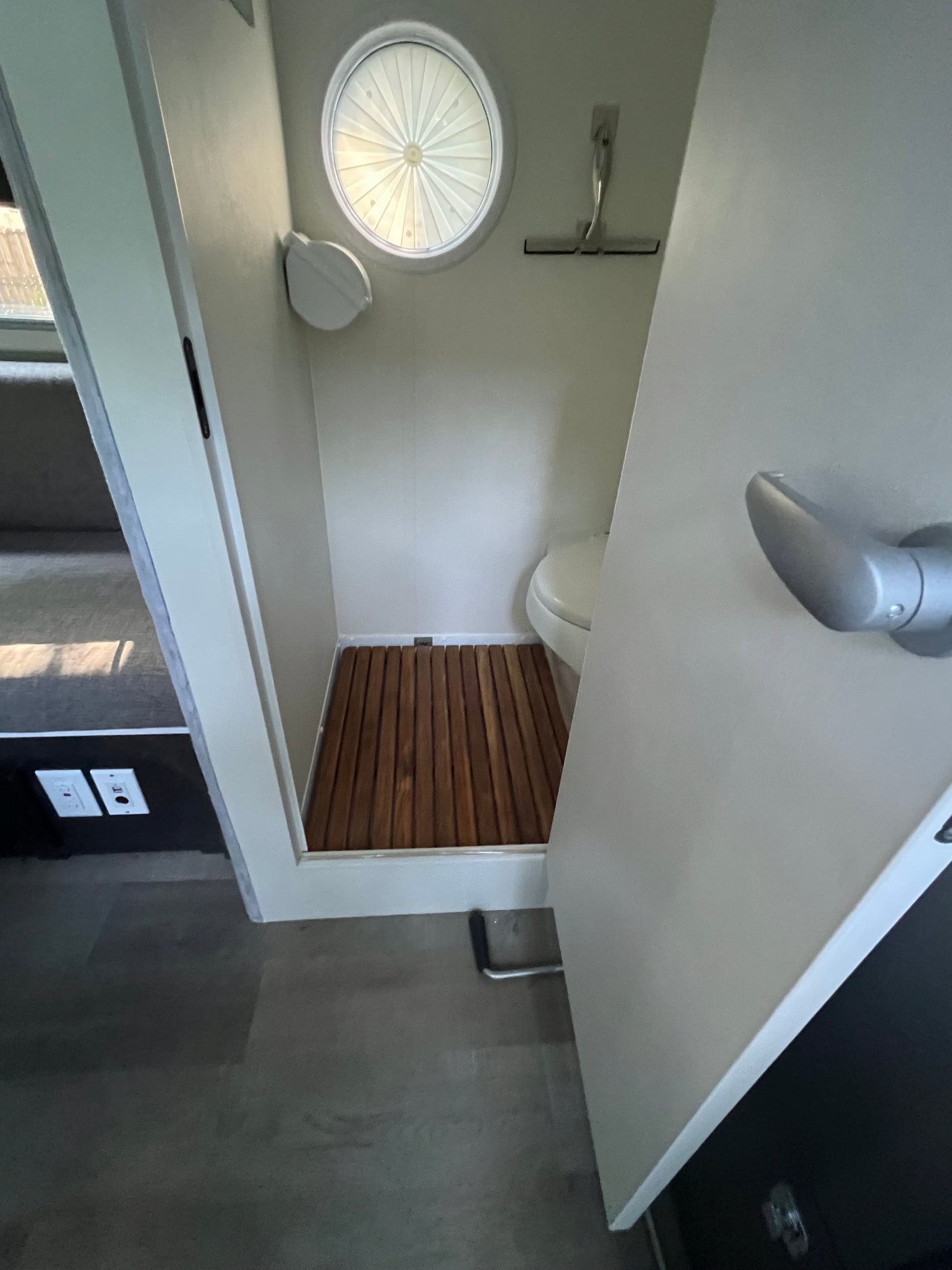 RV rental wet bath, showing compact toilet, wooden shower floor, and circular window with pleated shade.