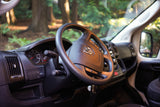 Driver's view inside an RV rental, showing the steering wheel, dashboard, and forest through the windshield.