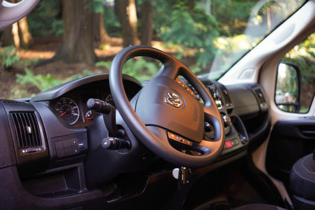 Driver's view inside an RV rental, showing the steering wheel, dashboard, and forest through the windshield.