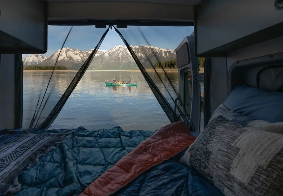 A cozy RV rental bed with a stunning view of a lake, snow-capped mountains, and a canoe carrying people.