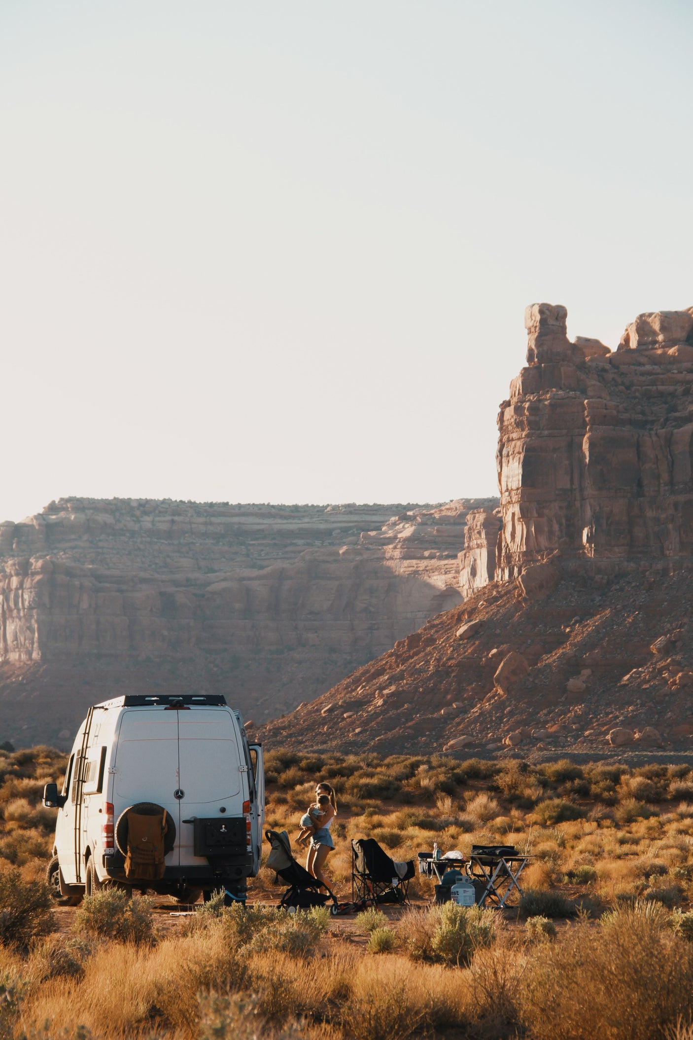 A white camper van (rv rental) is parked in a vast desert with towering red rock formations. A parent holds a child near camp chairs.