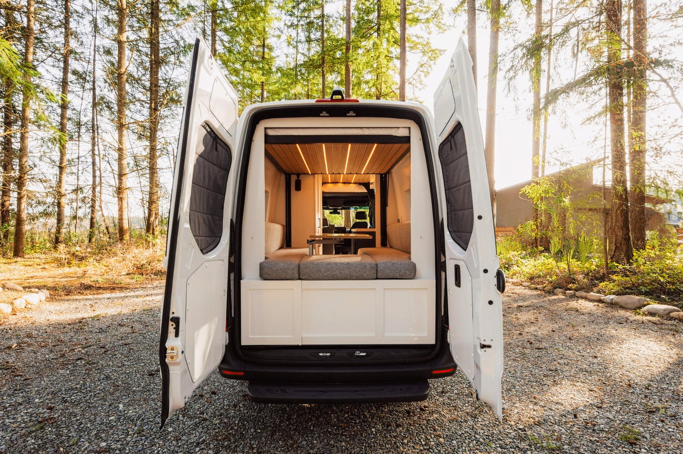 White camper van RV rental with back doors open, revealing a lit, wooden interior & grey cushions. Parked on gravel, surrounded by trees. — photo 9