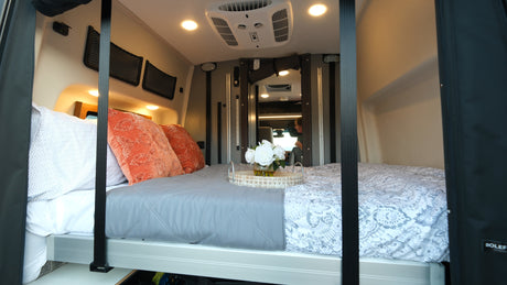 Comfortable bed inside an rv rental, with orange and patterned pillows, a floral tray, AC, and bright overhead lights. Ideal for travel.