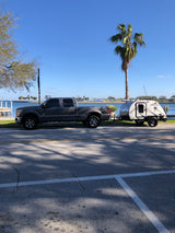 A gray pickup truck tows a teardrop RV by the sunny waterfront. Great for an rv rental trip, with palm trees visible.