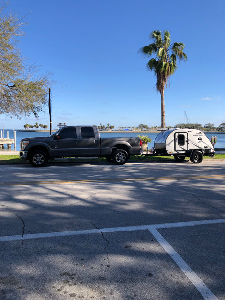 A gray pickup truck tows a teardrop RV by the sunny waterfront. Great for an rv rental trip, with palm trees visible.