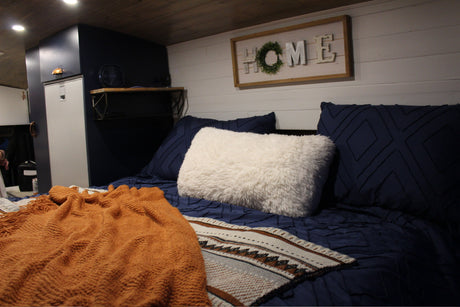 Cozy RV rental bedroom with navy blue bedding, a fluffy white pillow, orange blanket, and a HOME sign.