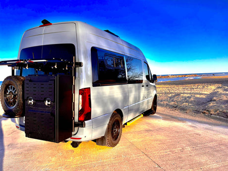 Custom light-grey camper van RV rental, with off-road tires & rear storage, parked on a sunlit beach under blue sky.