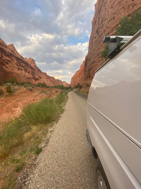 White rv rental van parked on a gravel road in a striking red rock canyon, winding through desert landscape under a cloudy sky.