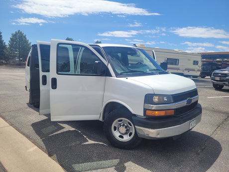 A white van with open doors is parked; a large RV rental and other vehicles are visible in the sunny background.
