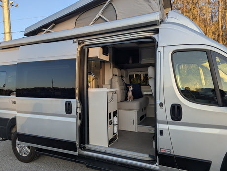Silver camper van for rv rental, door open revealing a cozy interior with a dog on the bench. Features a pop-top roof.