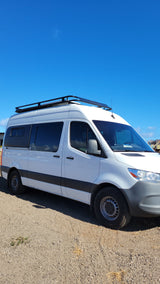 White Mercedes-Benz Sprinter van conversion with a roof rack, ready for your RV rental adventure. Parked on gravel.