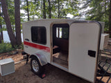This white Runaway Navigator travel trailer, an rv rental, is lakeside with its door open, offering a cozy retreat in nature.