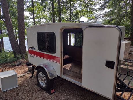 This white Runaway Navigator travel trailer, an rv rental, is lakeside with its door open, offering a cozy retreat in nature.