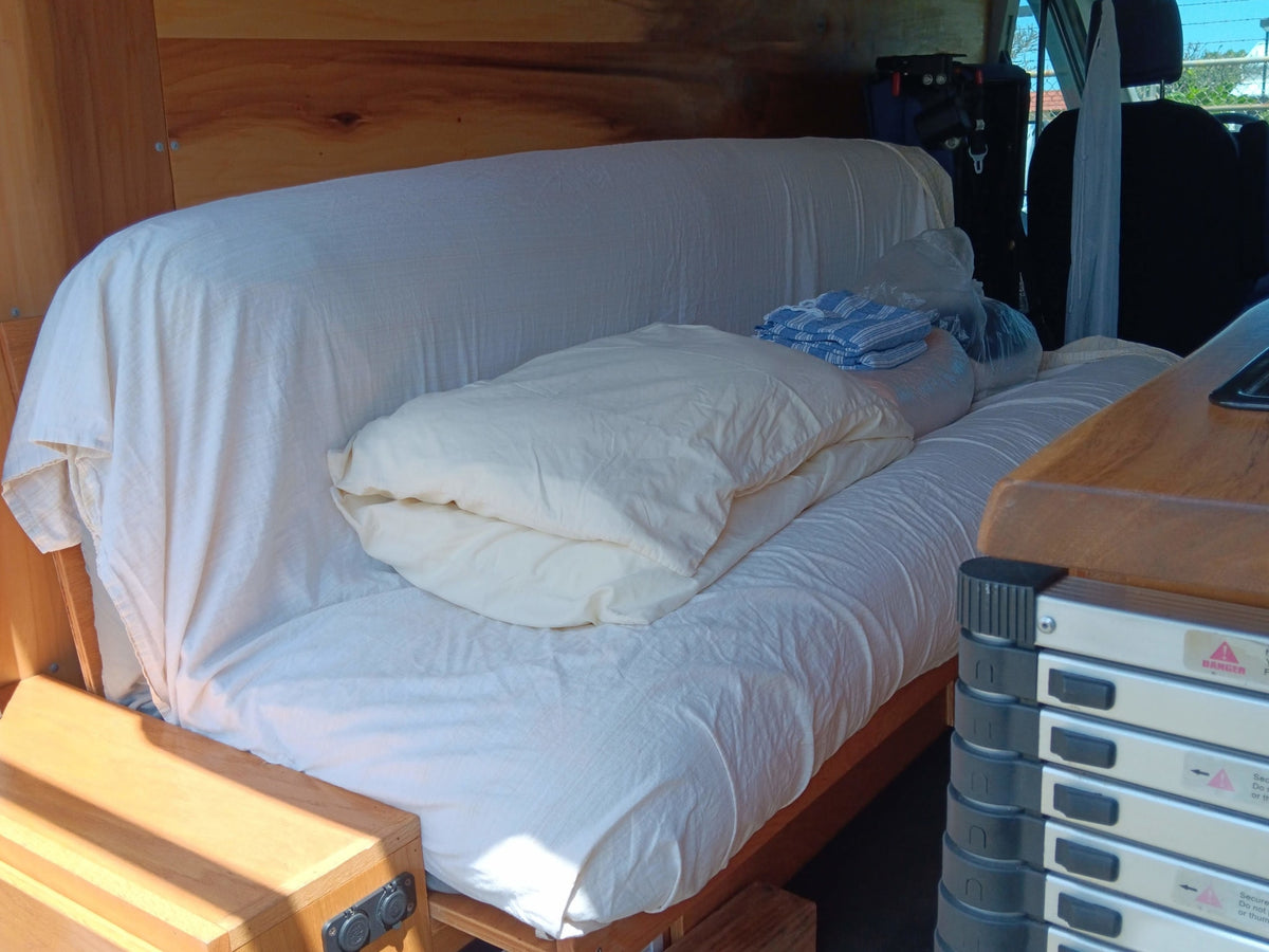 RV rental interior: custom wooden build with a comfortable bench/bed setup, white bedding, and travel essentials. — photo 11