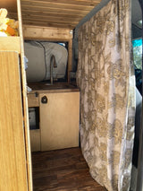 Inside an RV rental: A camper van's kitchen area with a sink, modern faucet, wood paneling, and a decorative privacy curtain.