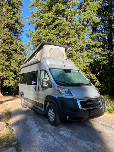 This silver pop-top RV rental, a camper van, is parked in a sunny wooded campsite, ready for adventure.