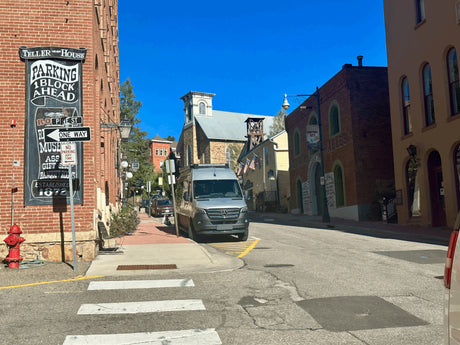 A silver RV rental van is parked on a vibrant historic street with brick buildings under a clear blue sky in Central City.