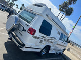 White Pleasure-Way RV rental, featuring a spare tire & graphics, parked on a sunny day near a beach and pier.